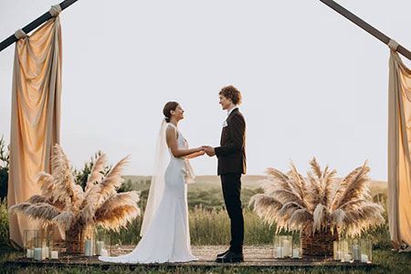 A couple holds hands during an outdoor wedding ceremony under a decorative arch with draped fabric and pampas grass.