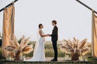 A couple holds hands during an outdoor wedding ceremony under a decorative arch with draped fabric and pampas grass.