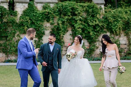 A wedding party walks across a lawn in front of an ivy-covered stone wall, smiling and conversing.