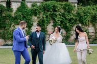 A wedding party walks across a lawn in front of an ivy-covered stone wall, smiling and conversing.