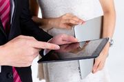 A business professional in a suit and striped tie points at a tablet held by a colleague in a white textured dress.