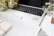 Two gold wedding rings rest on a laptop keyboard surrounded by calendars, small gift boxes, and sprigs of baby's breath.