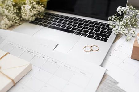 Two gold wedding rings rest on a laptop keyboard surrounded by calendars, small gift boxes, and sprigs of baby's breath.