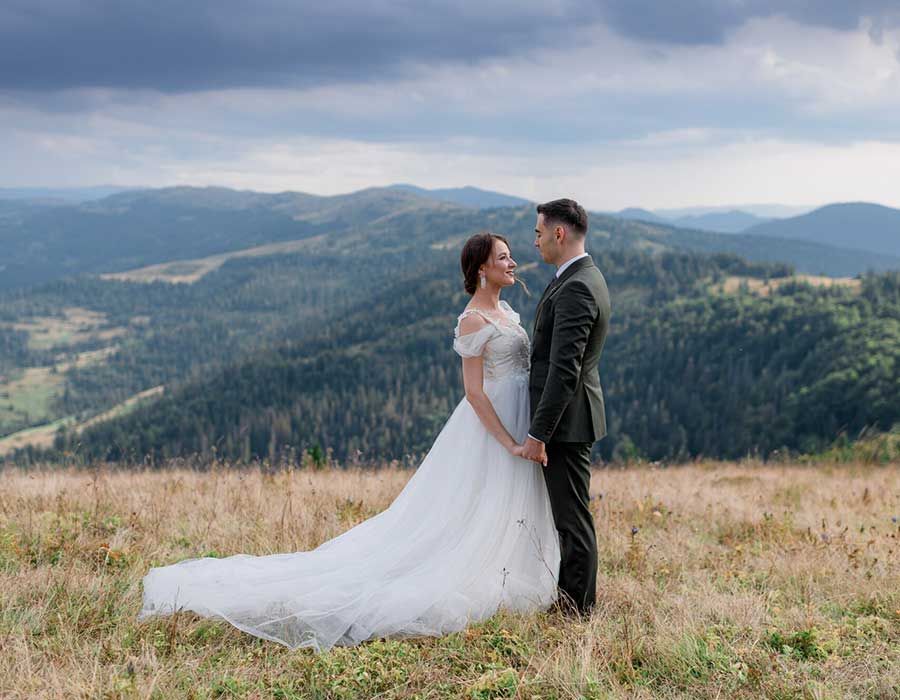 A couple in wedding attire holding hands while facing each other on a grassy mountain hillside under a cloudy sky.