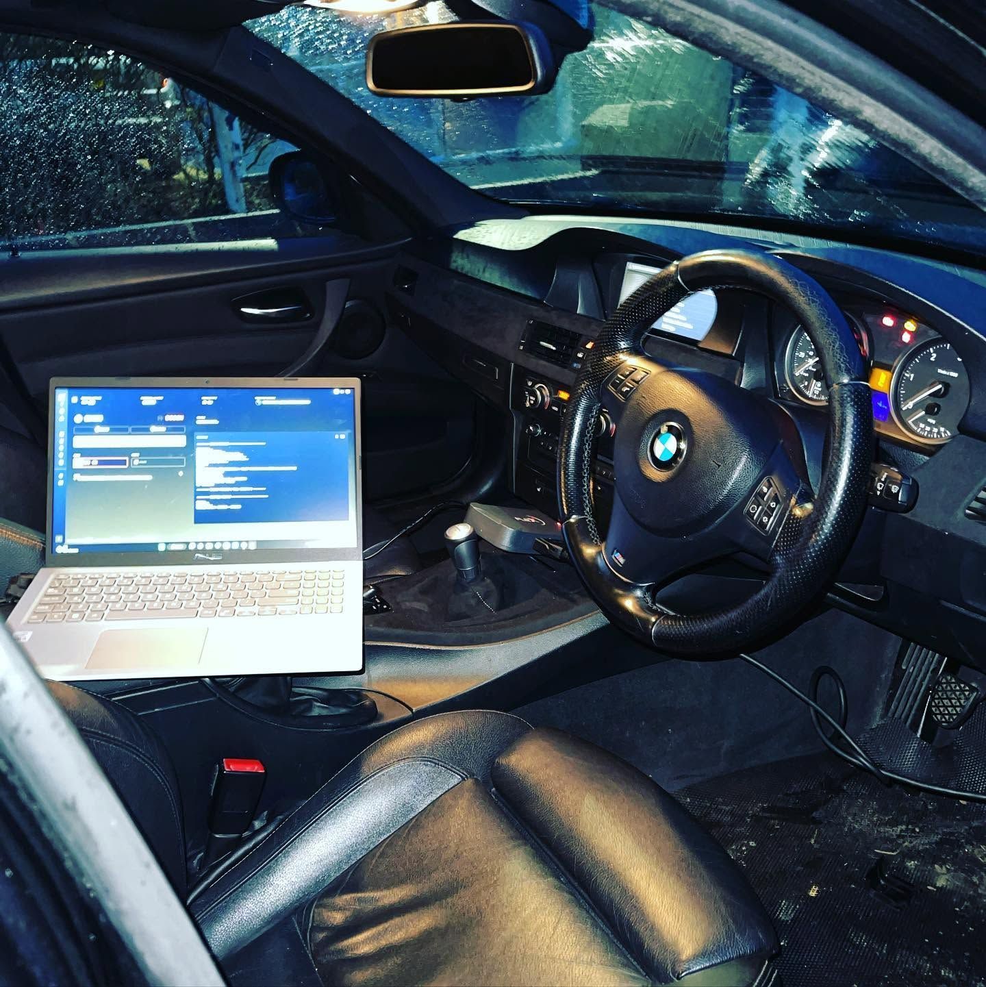 Laptop open on a BMW driver’s seat inside a car, with the dashboard and steering wheel visible.