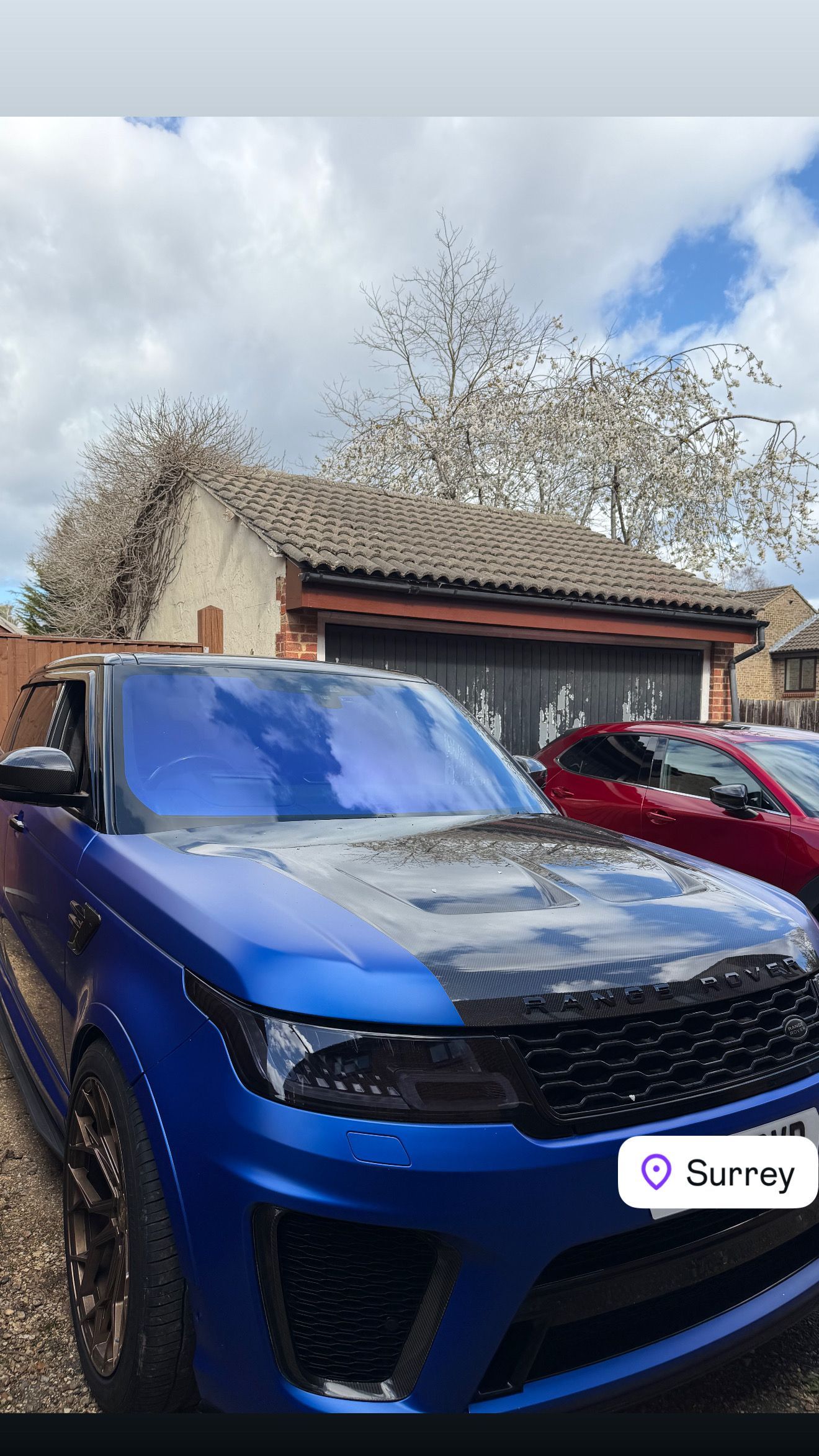 Blue SUV parked in a driveway beside a red car, in front of a house under a cloudy sky.