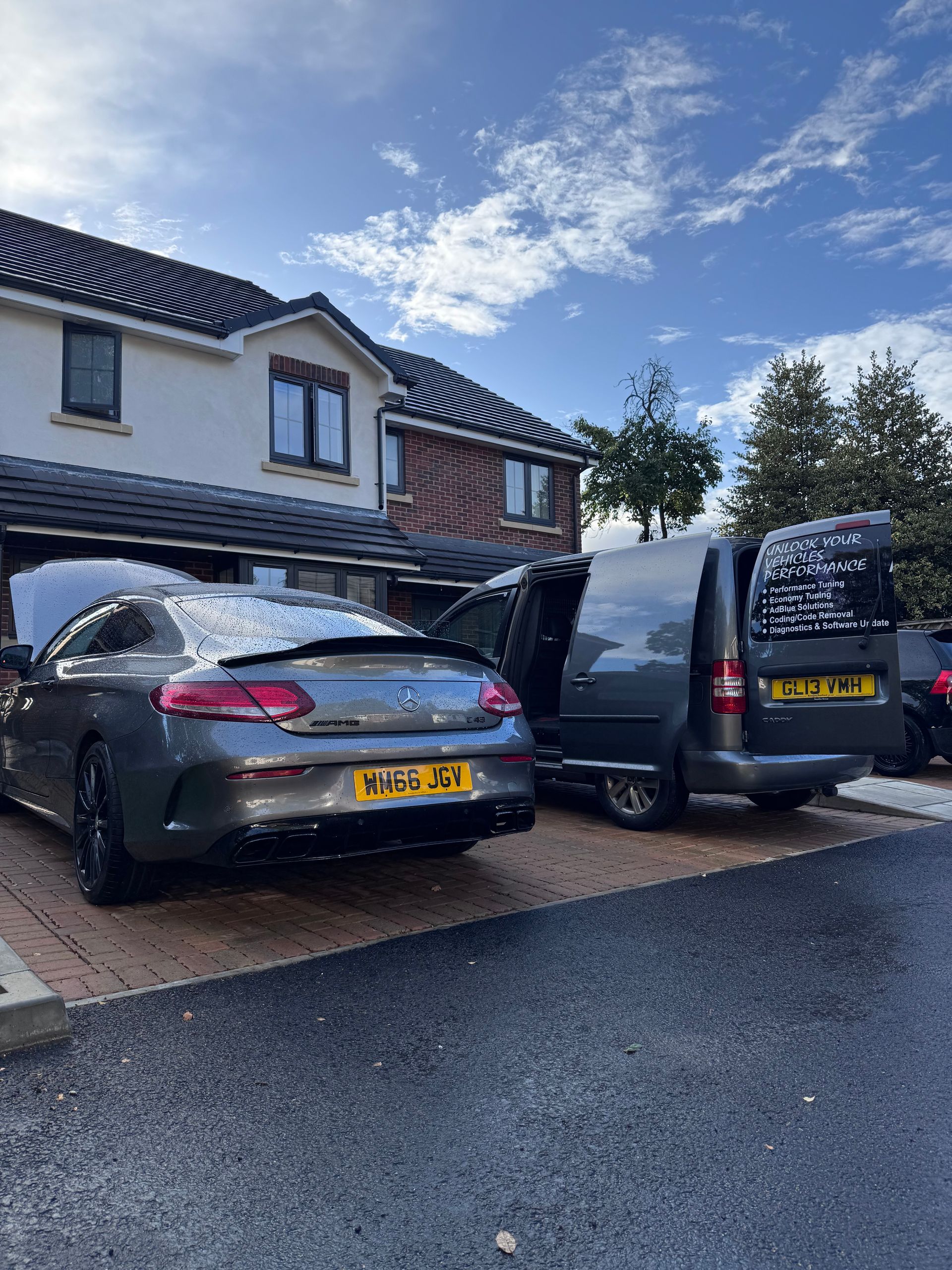 Two parked cars, a gray sedan and black van, in a driveway beside a house under a blue sky.