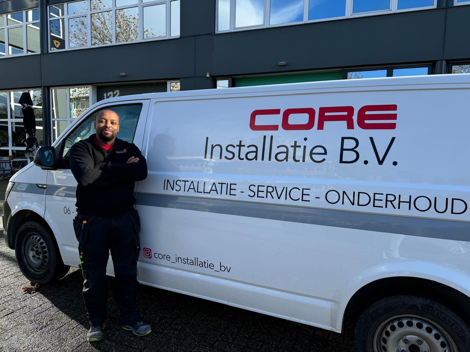 Een man in werkuniform staat naast een witte CORE-bus met zijn armen over elkaar. Buiten, voor het gebouw.