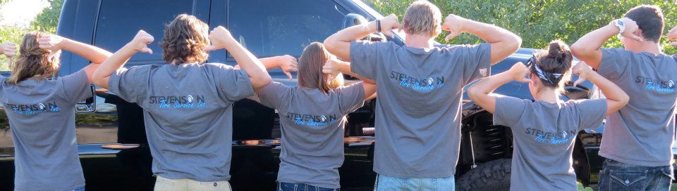 Group of people in grey t-shirts with arms flexed overhead, posing in front of a dark vehicle.