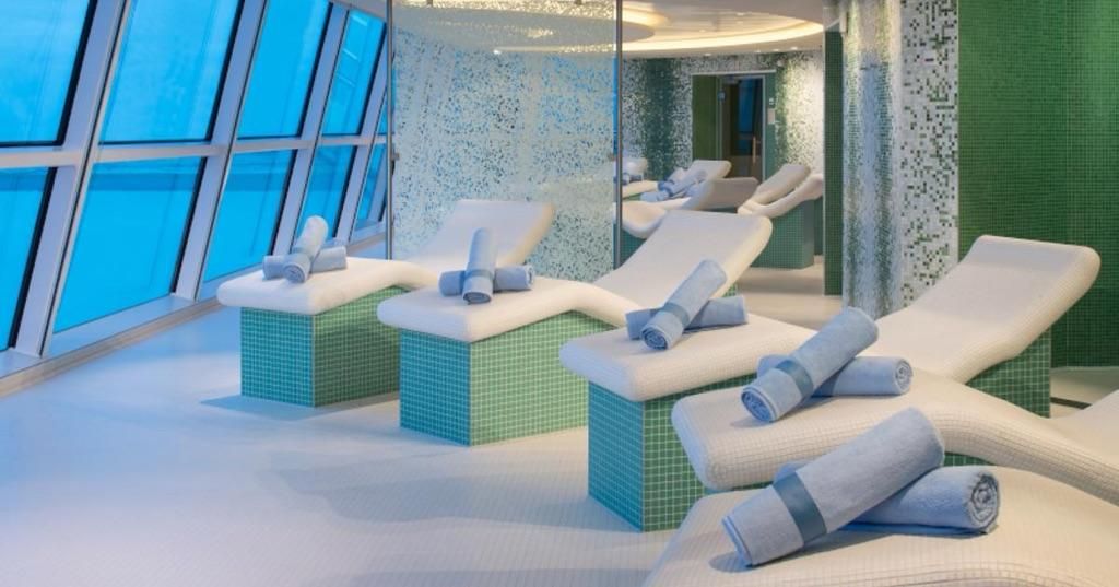 Luxury spa lounge with white loungers, blue cushions, and floor-to-ceiling windows overlooking water