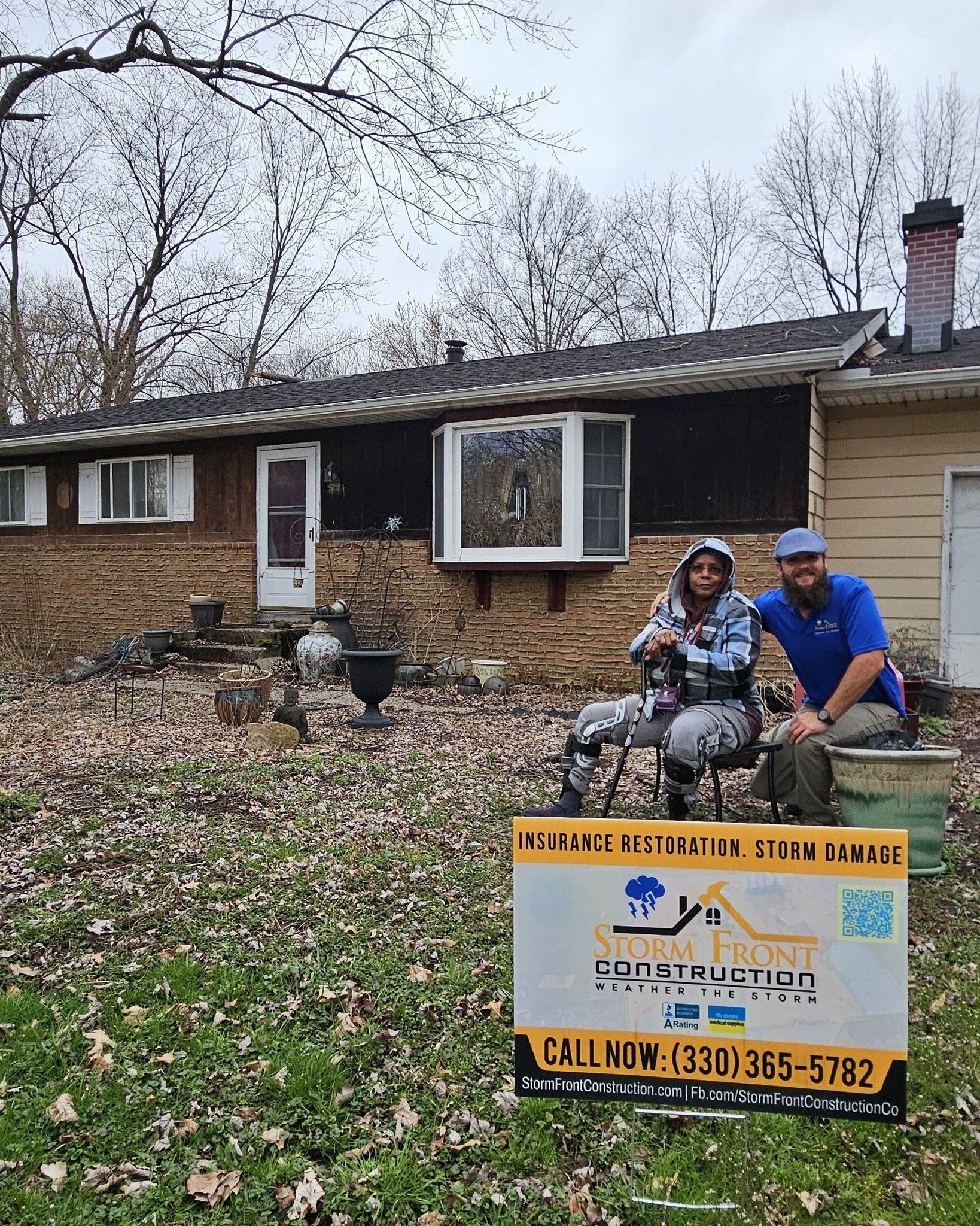 Two people sitting on a lawn in front of a brick house with a yellow sign in the foreground