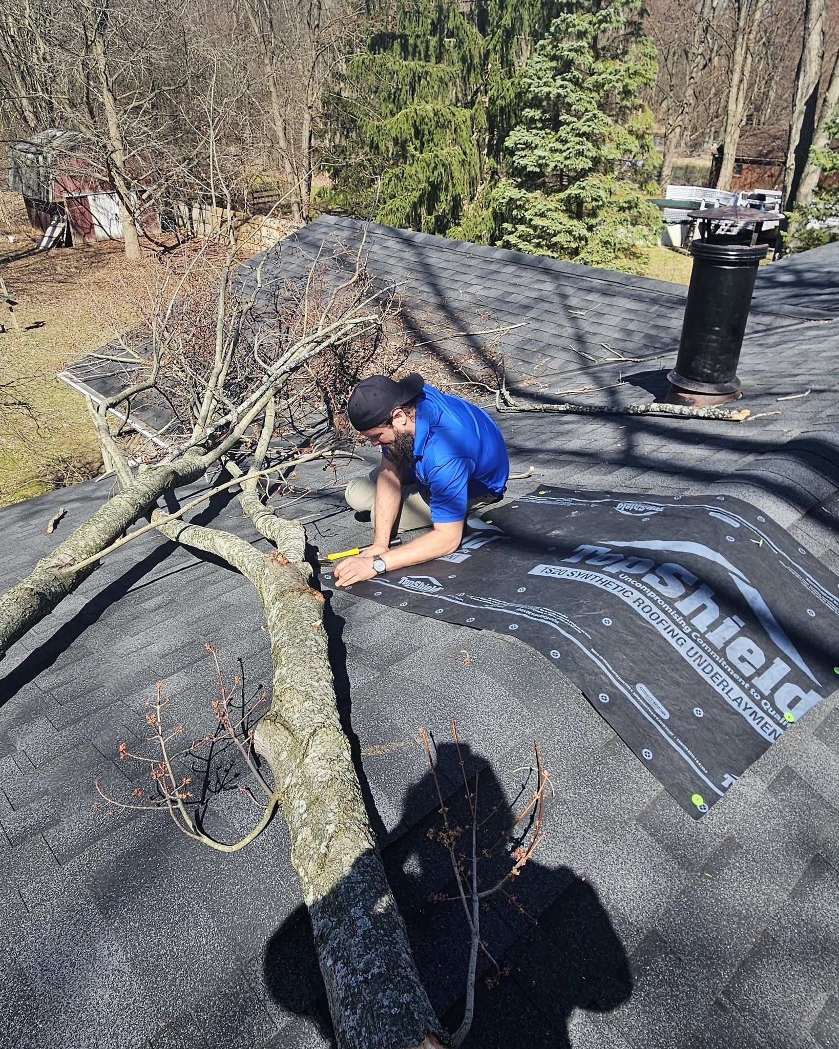 Person kneels on a shingled roof, repairing damaged underlayment near a chimney.