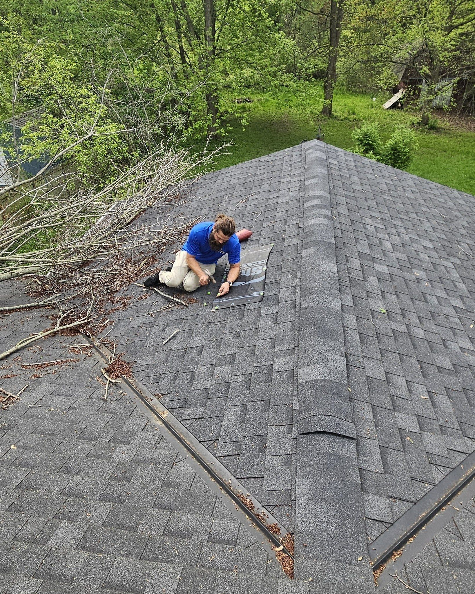 Person in a blue shirt crawling on a steep gray shingled roof surrounded by trees.