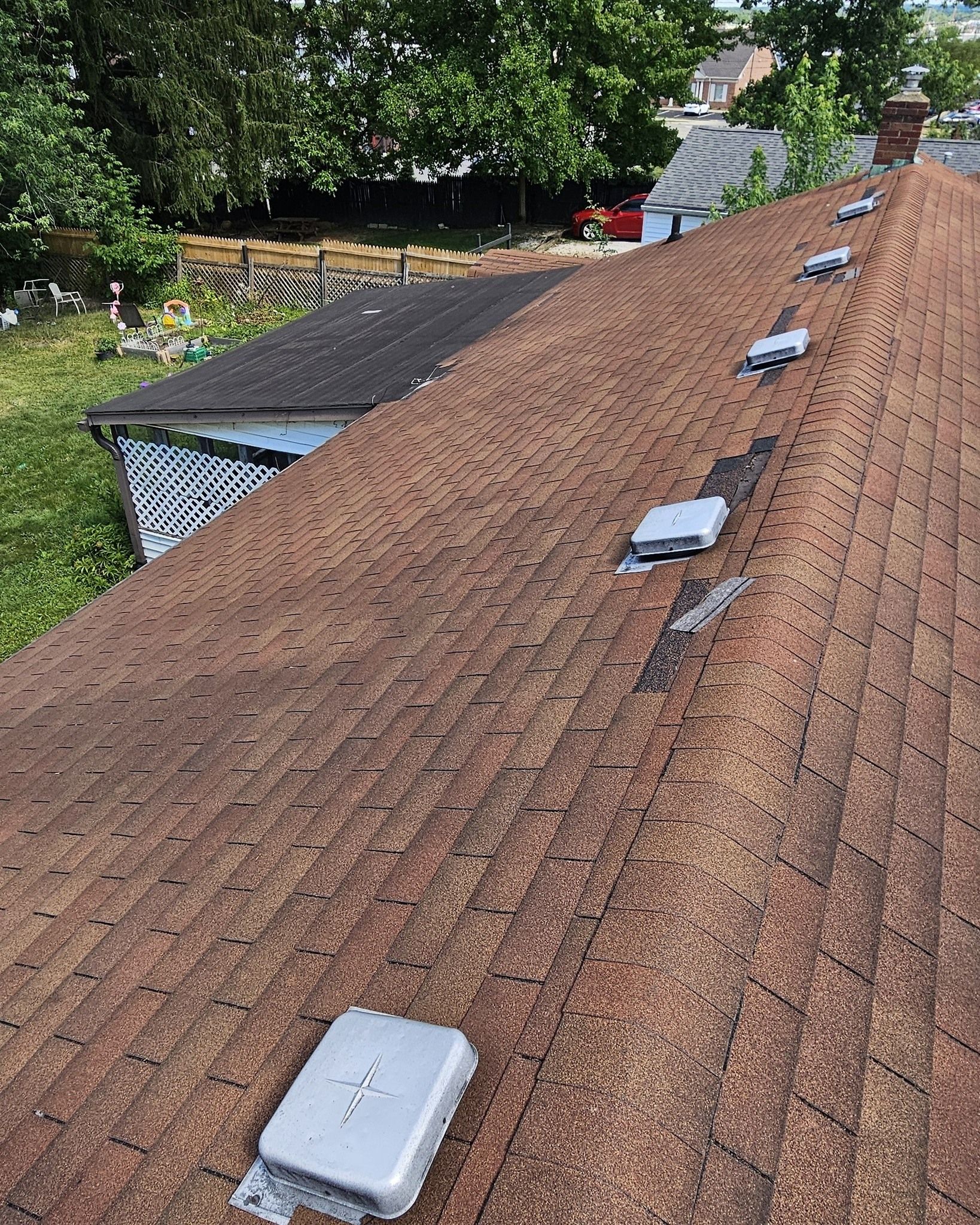 Reddish-brown shingled roof with several white roof vents near a wooded residential area