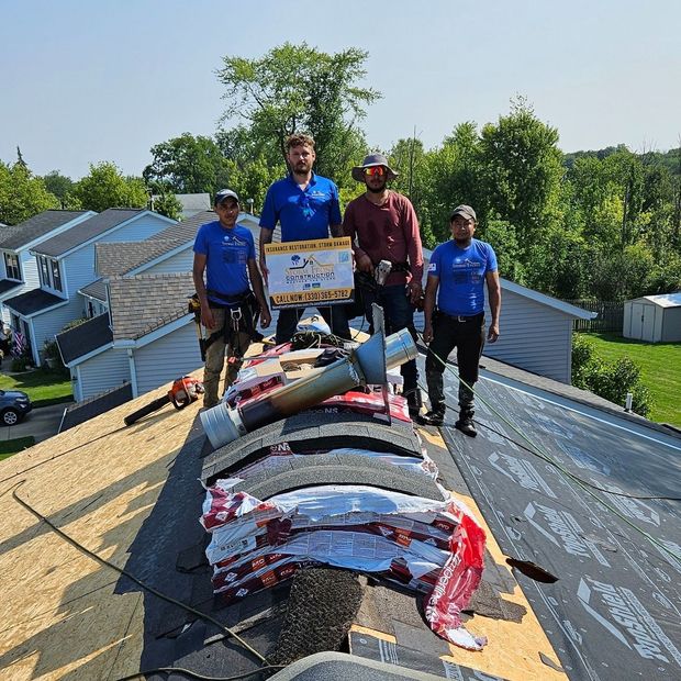 Four workers stand on a rooftop beside rolled roofing materials and a solar panel installation.