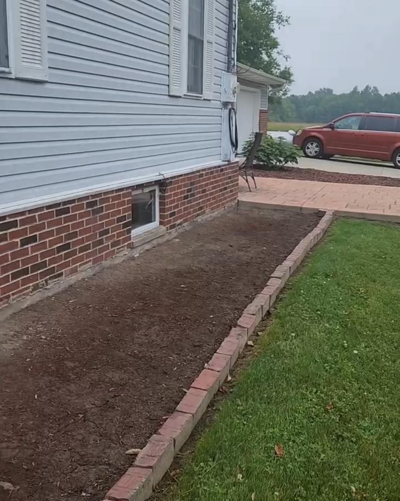 Fresh soil bed along a brick house beside a lawn, with a red car parked in the driveway.