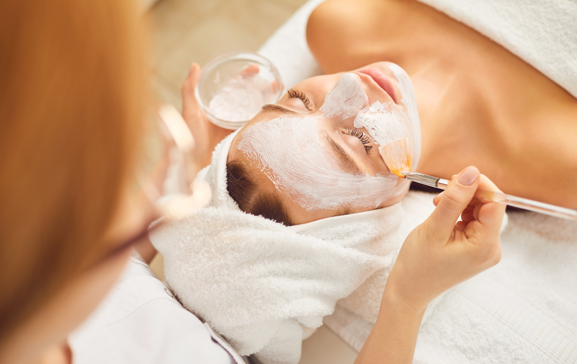 Esthetician applying a facial mask to a client lying on a spa bed with a towel on her head