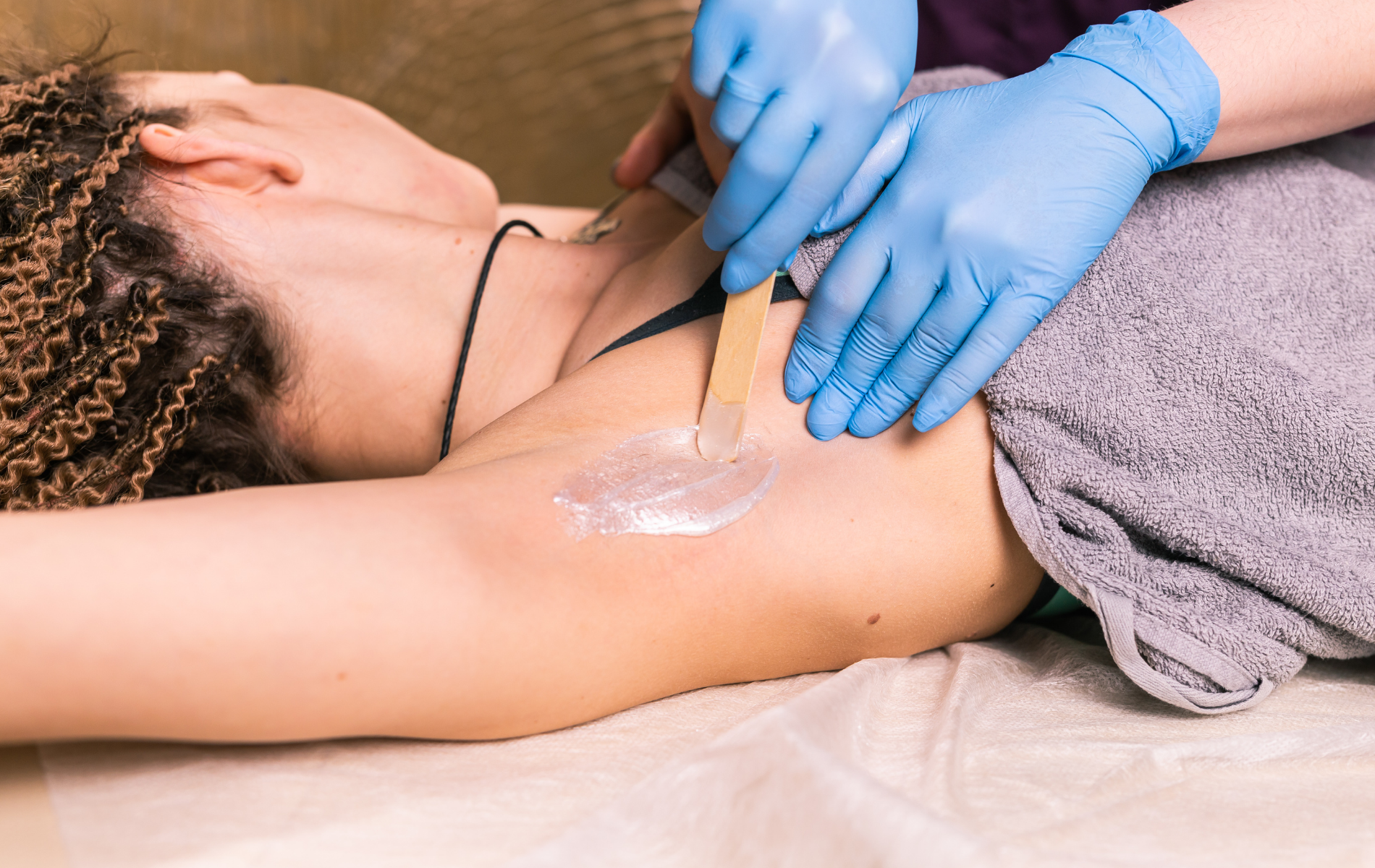 Gloved hands apply wax to an armpit during a waxing treatment on a spa bed