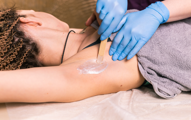 Gloved hands apply wax to a person’s underarm during a hair removal treatment.
