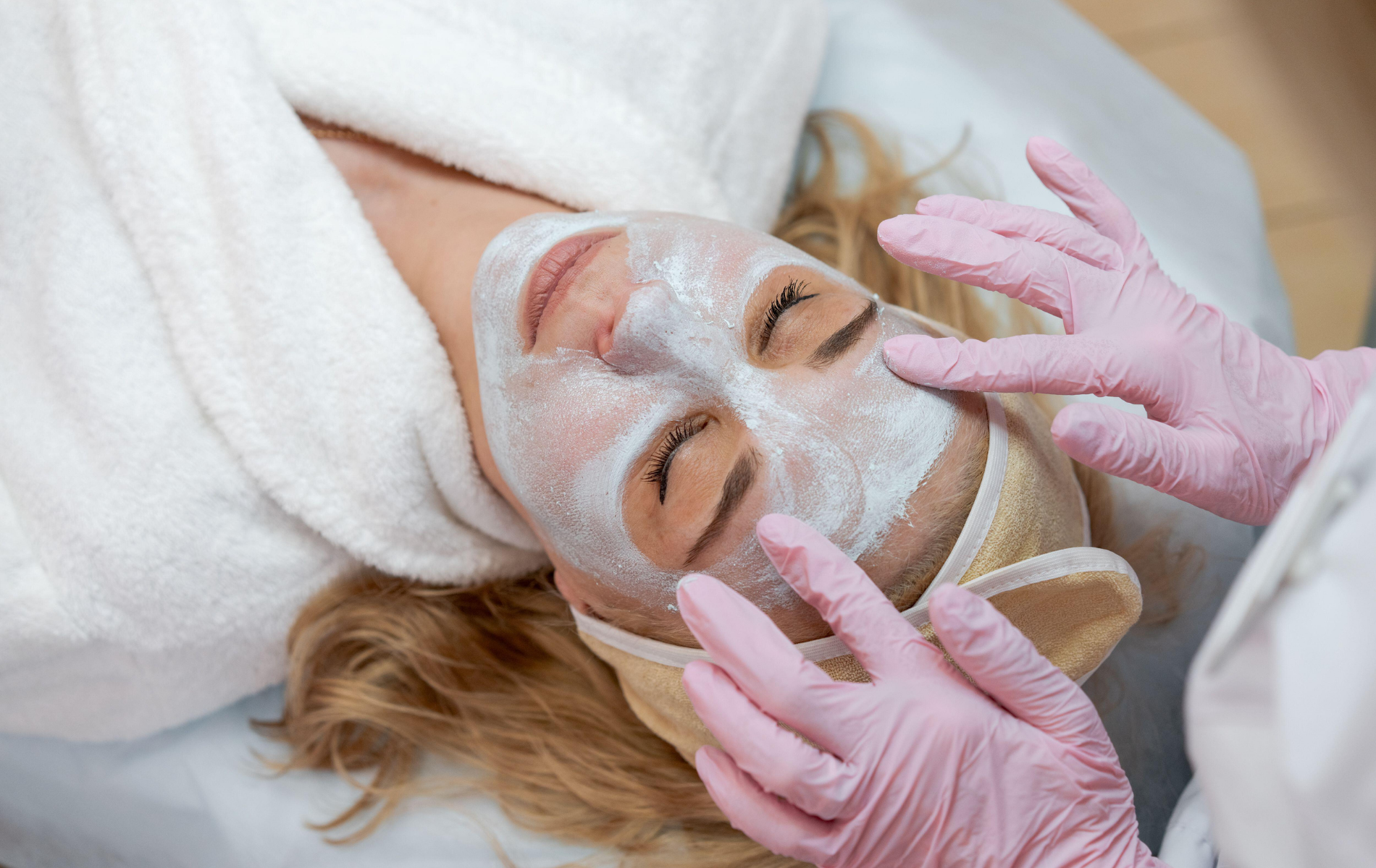 Woman receiving a facial treatment while a therapist applies a white mask with pink gloves