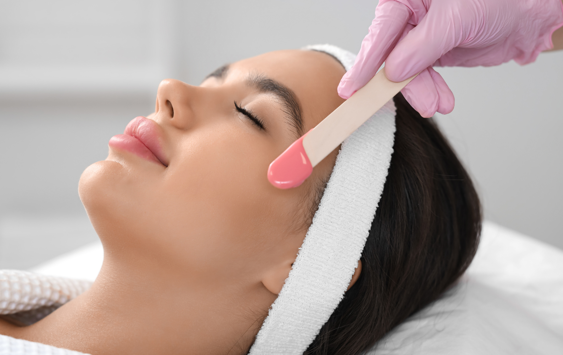 Esthetician applying pink wax to a client’s eyebrow during a facial treatment