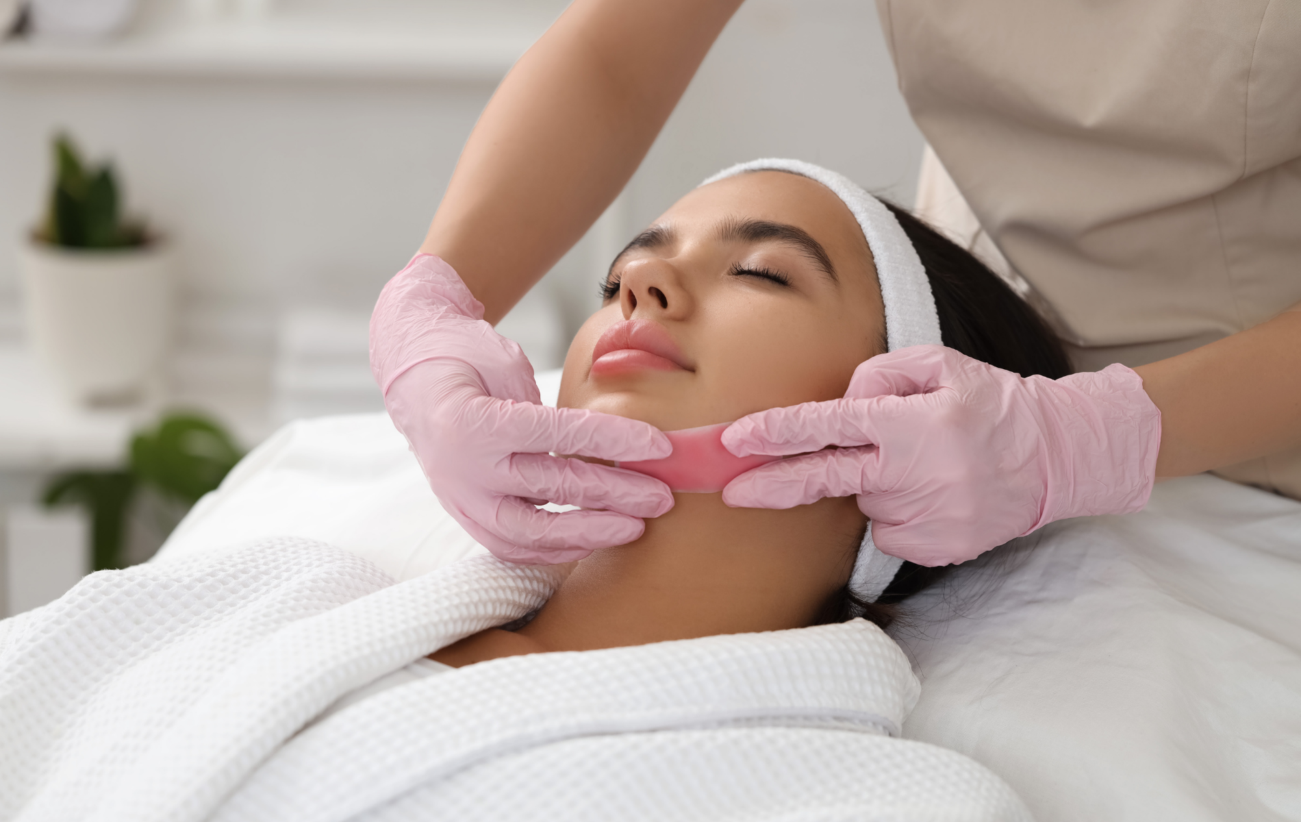 Facial treatment in progress on a woman lying on a spa bed, with pink-gloved hands massaging her chin.