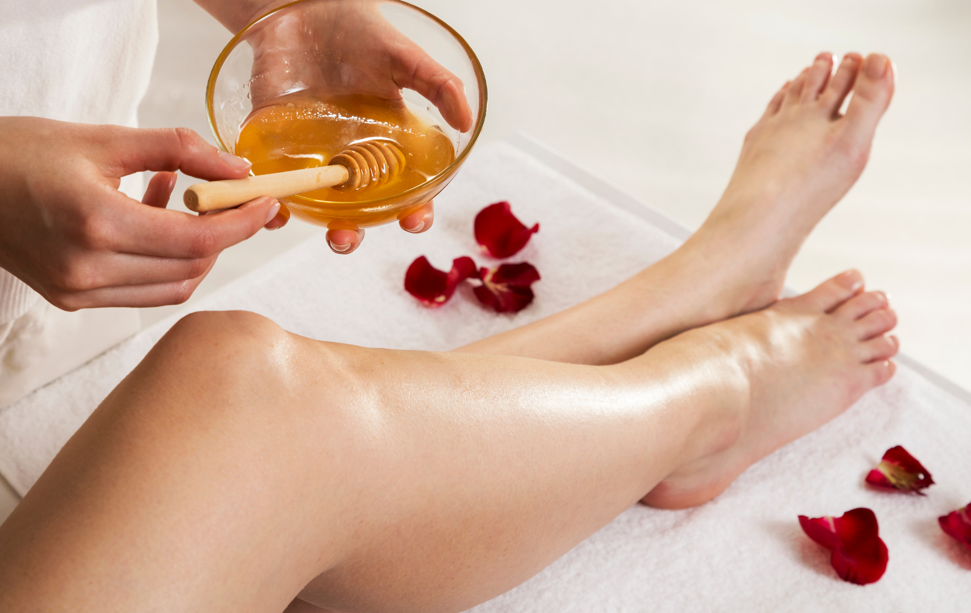 Legs receiving warm wax treatment with a spatula, white towel, and rose petals nearby