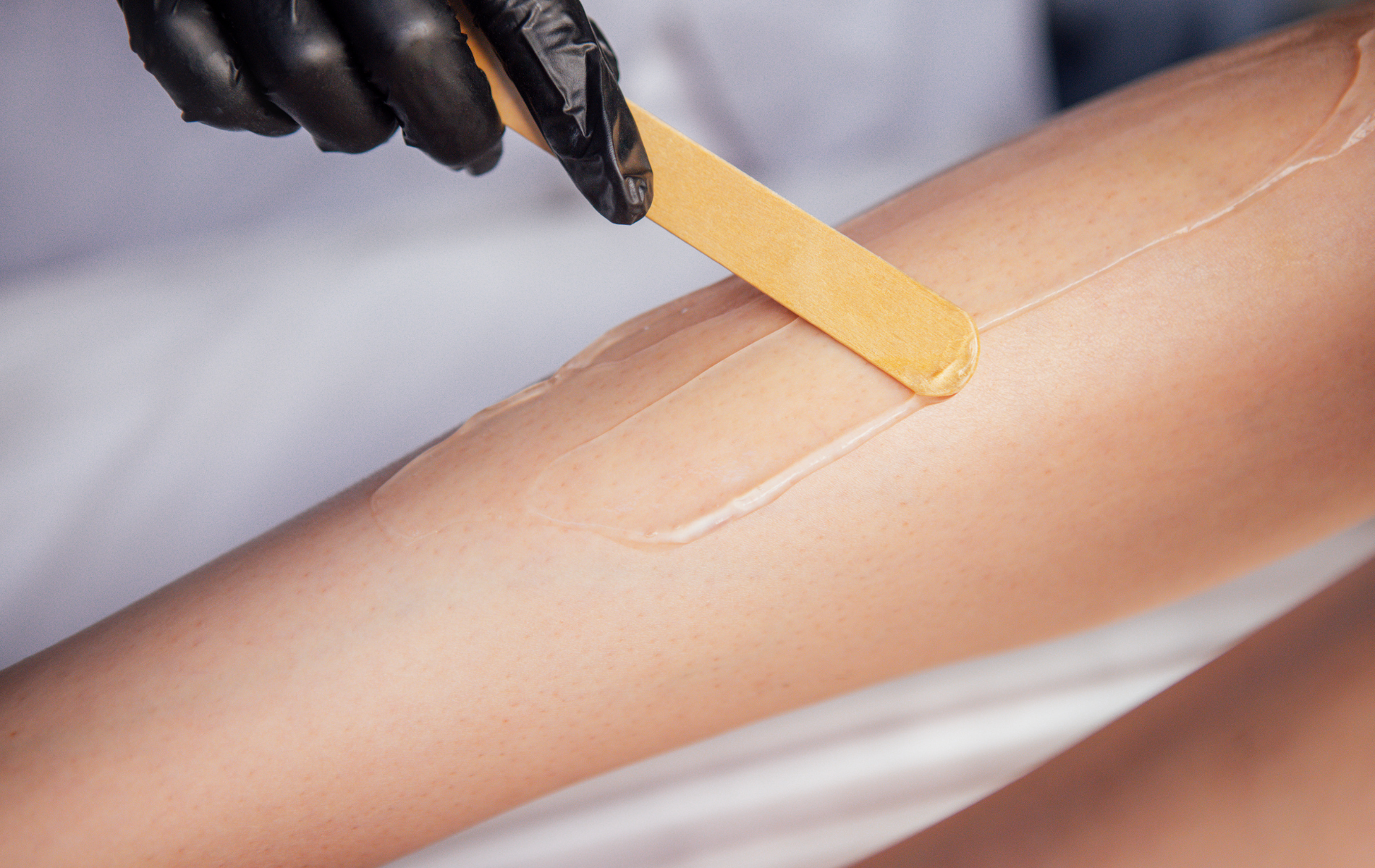 Gloved hand applying wax with a wooden spatula to a leg during hair removal treatment