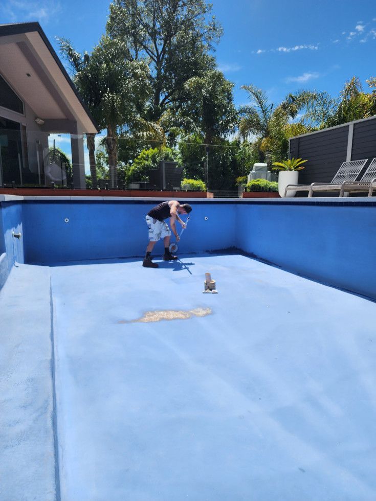Person cleaning a drained, blue swimming pool outdoors on a sunny day.