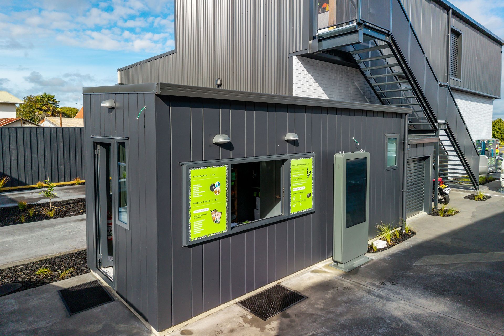 Exterior of a dark gray drive-thru kiosk with a digital menu board.  Building has a metal exterior and an attached staircase.