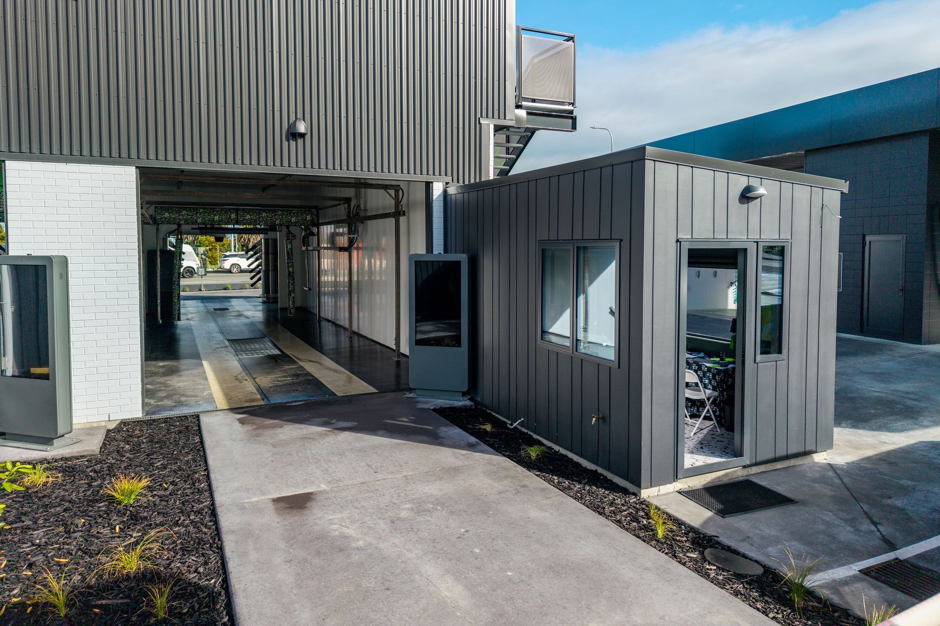 Exterior of a car wash facility with a booth. Gray and black building with a drive-through lane.
