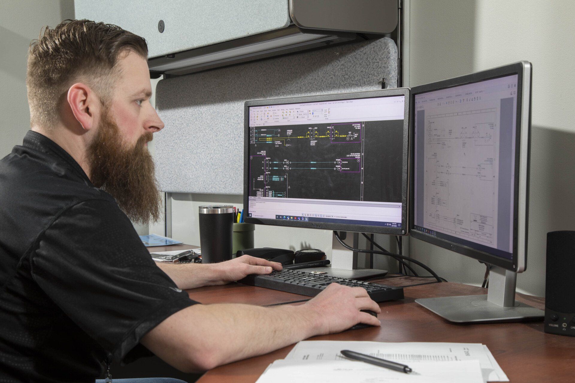 A man with a beard is sitting at a desk in front of two computer monitors.