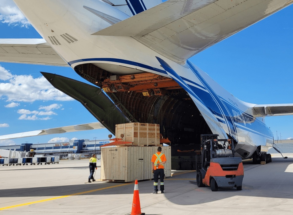 Cargo being loaded into an open airplane cargo hold on the tarmac, with workers and a forklift.