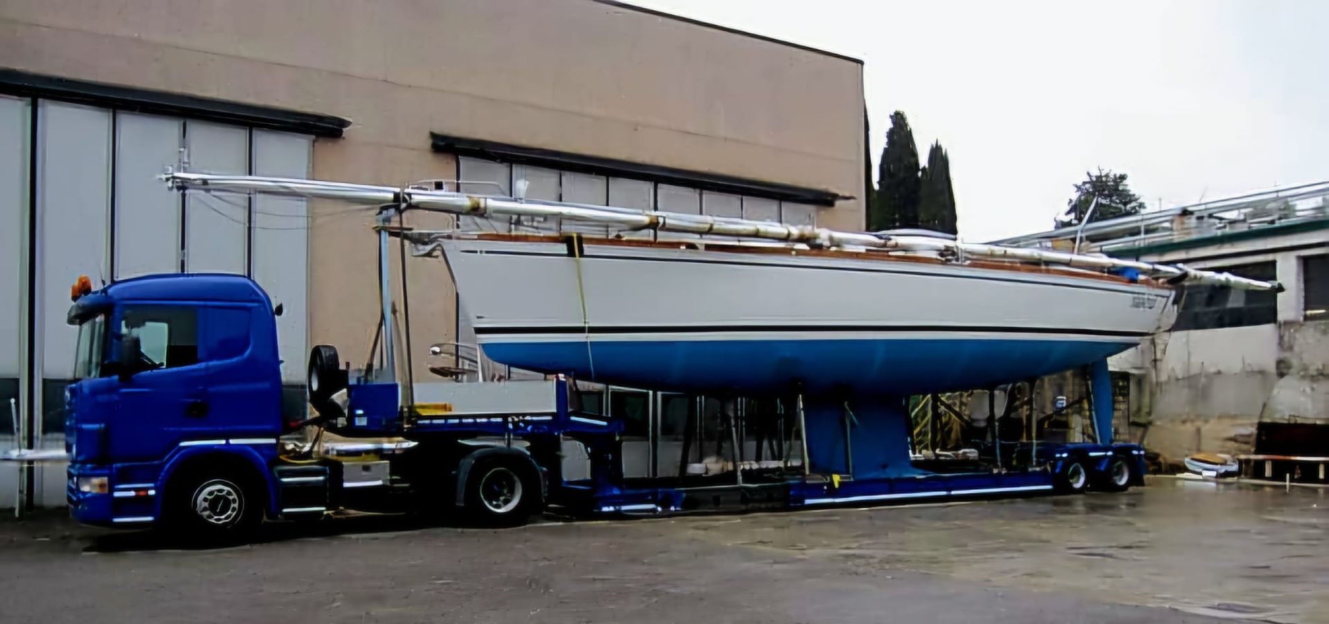 Blue truck hauling a large sailboat on a trailer in an industrial lot