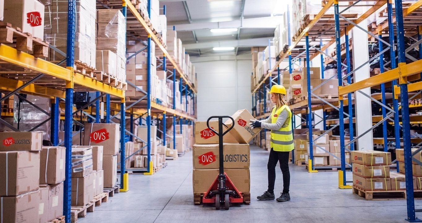 Warehouse worker with pallet jack beside stacked boxes in a bright storage aisle