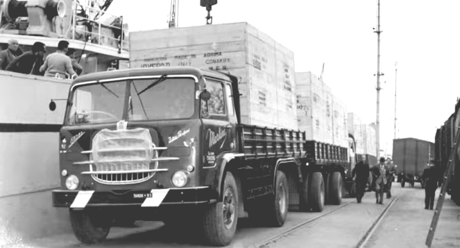 Vintage cargo truck on a dock beside a ship, with stacked crates and workers in the background.
