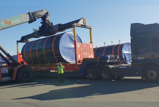 Crane loading a large wrapped cylindrical tank onto a flatbed truck in an industrial yard