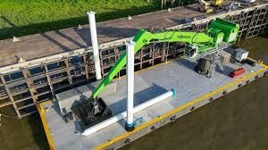 Aerial view of a green excavator on a flatbed barge beside a dock and tall white pole