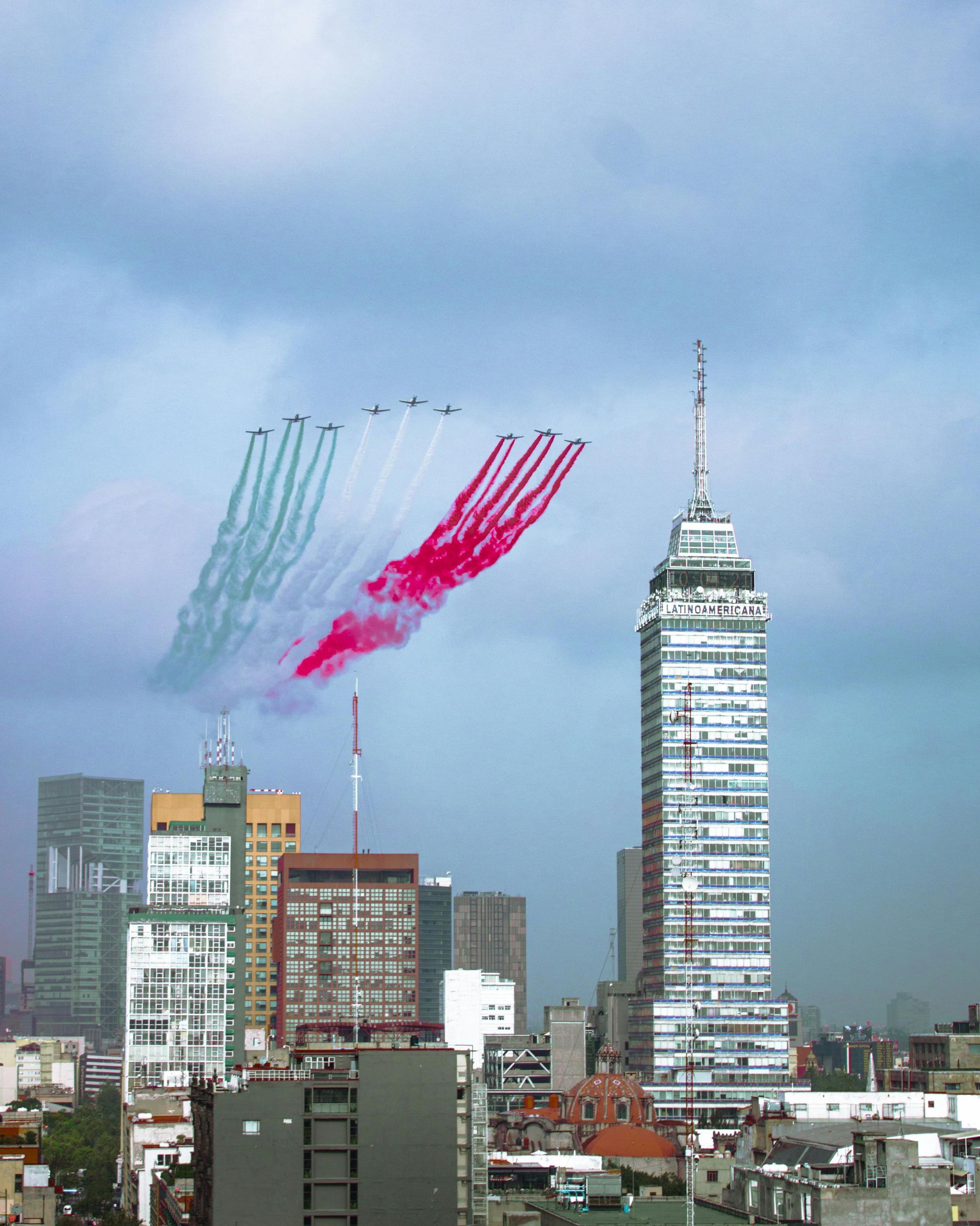 Jet planes trail teal and red smoke above a city skyline, beside a tall tower.