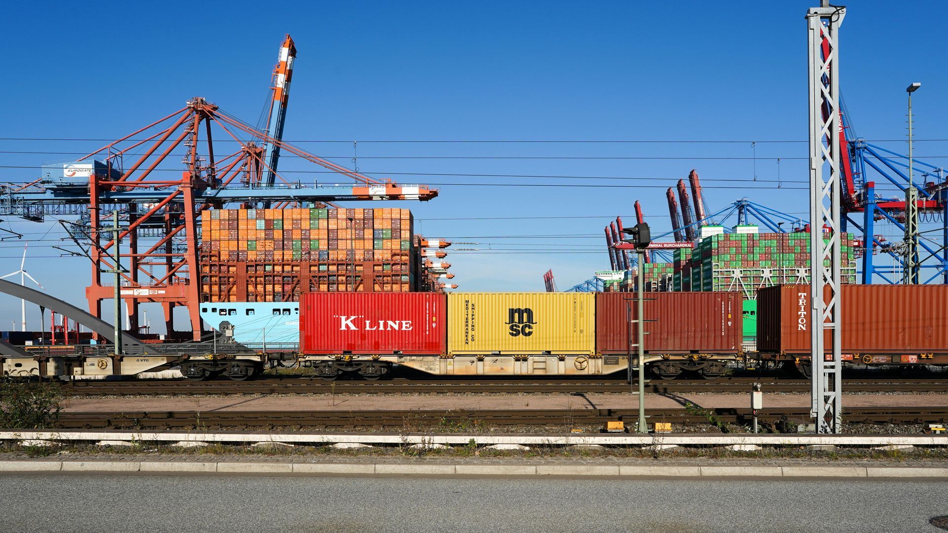 Container train at a busy shipping port with cranes and stacked cargo containers under a clear blue sky