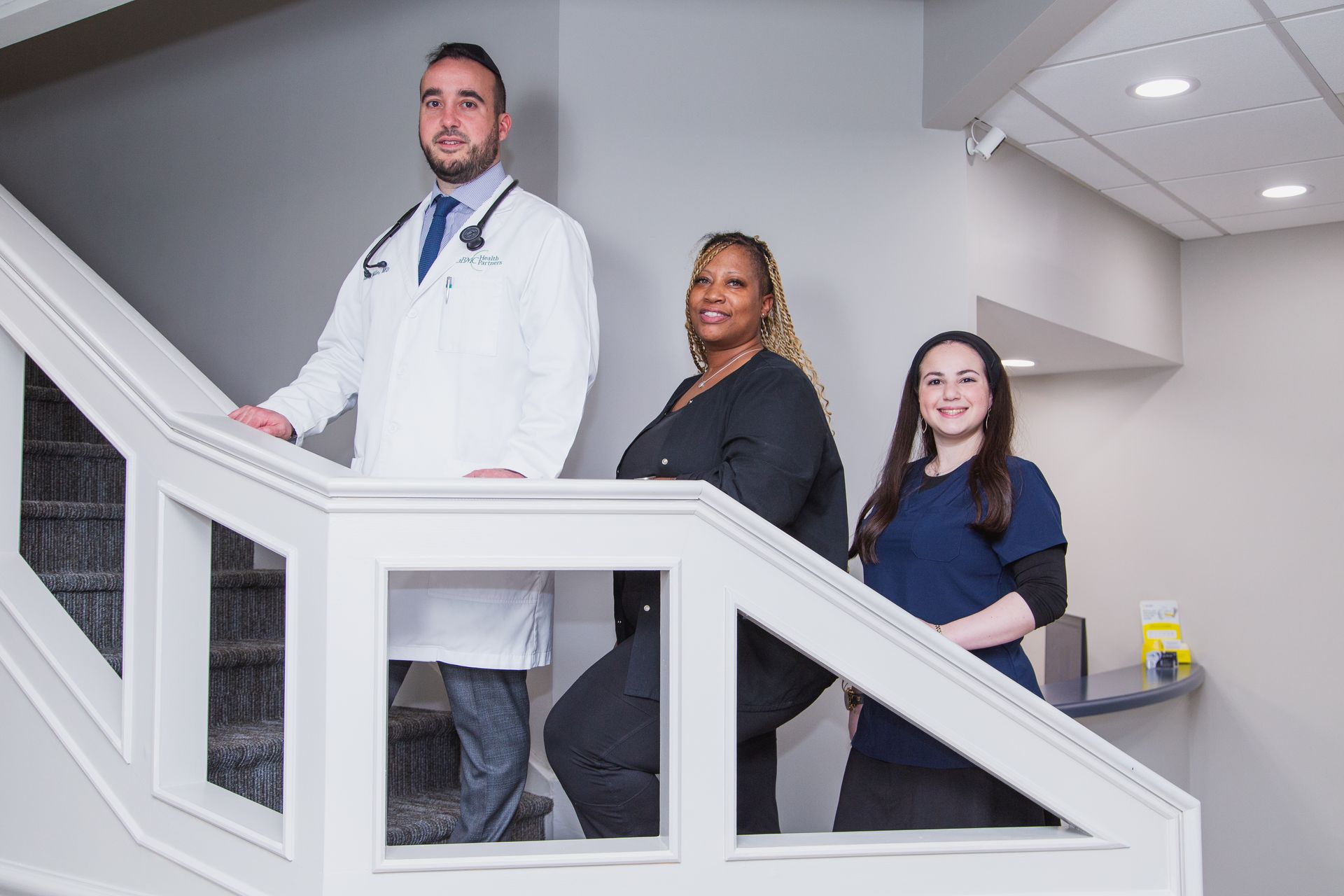 Three medical staff standing on a staircase in a clinic hallway, wearing white and navy uniforms.