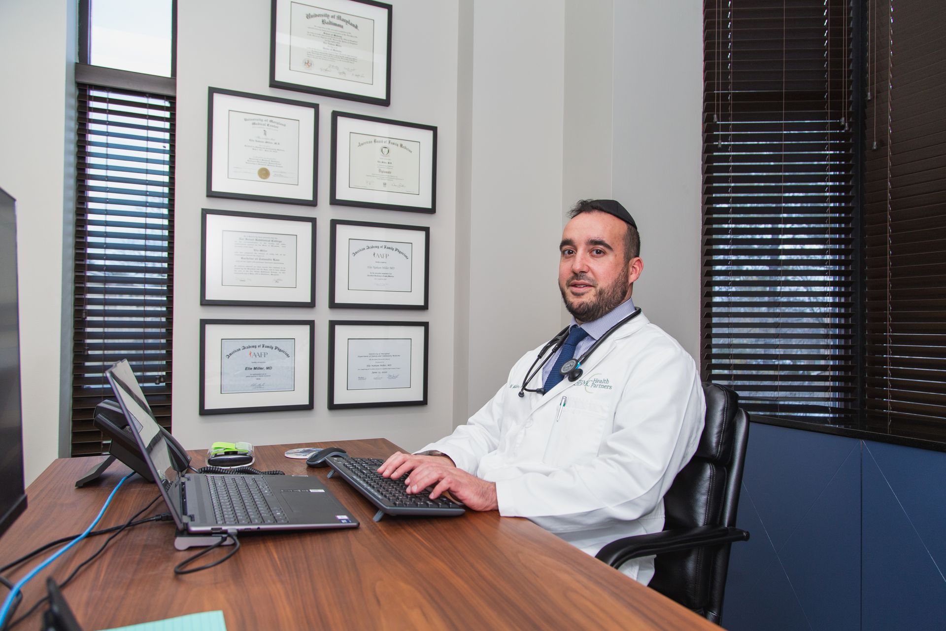 Doctor seated at a desk in an office, using a laptop with framed certificates on the wall.