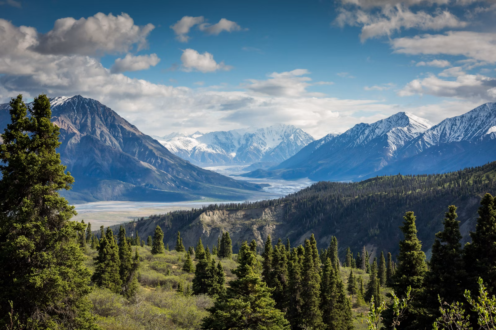 Snowy mountain range above a forest under a blue sky with scattered clouds