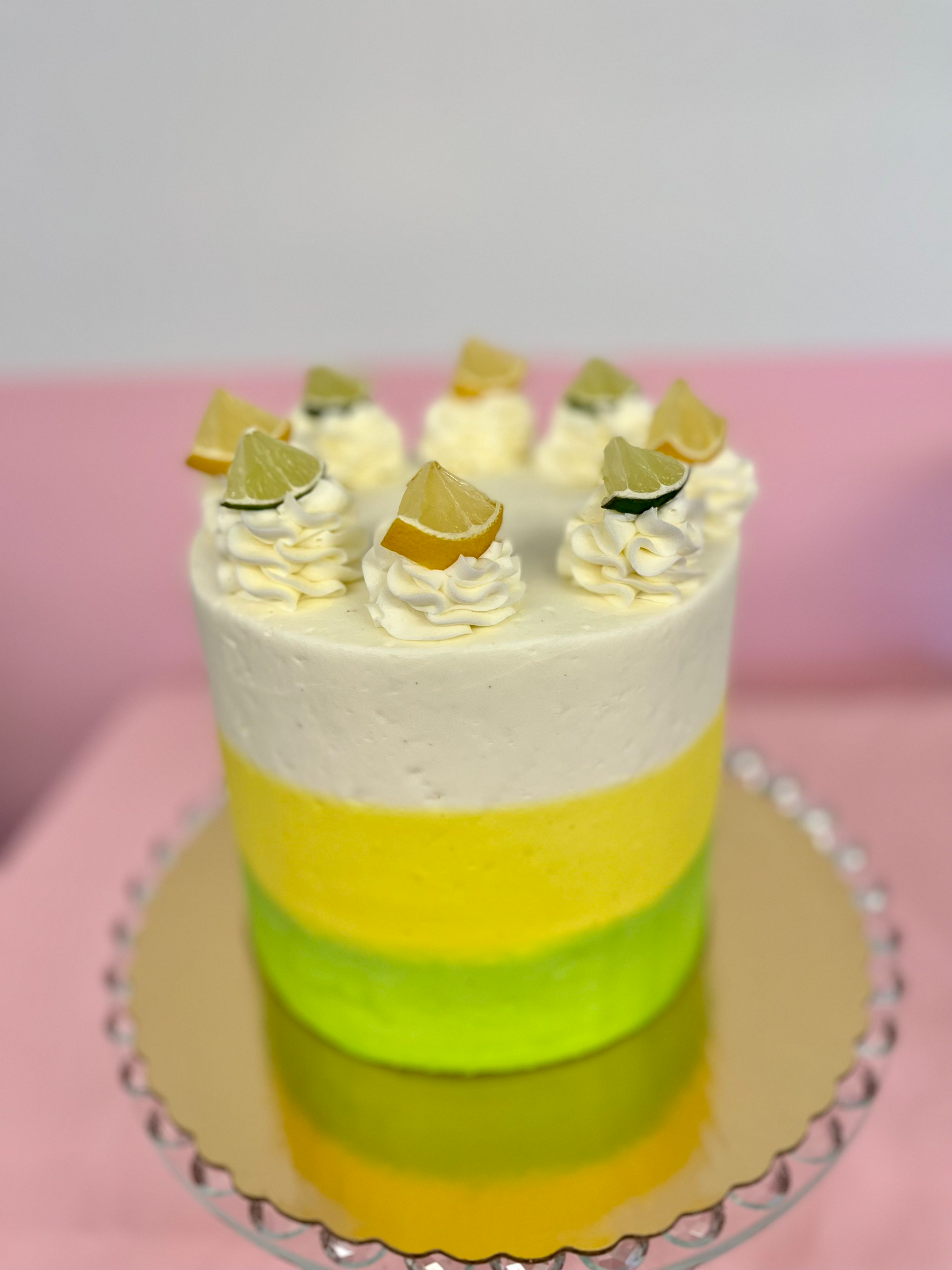 A yellow , green and white cake with lemon slices on top is on a glass plate on a table.