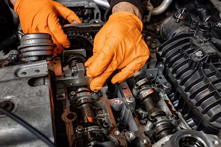 Gloved hands repairing a car engine with exposed valve train and orange work gloves