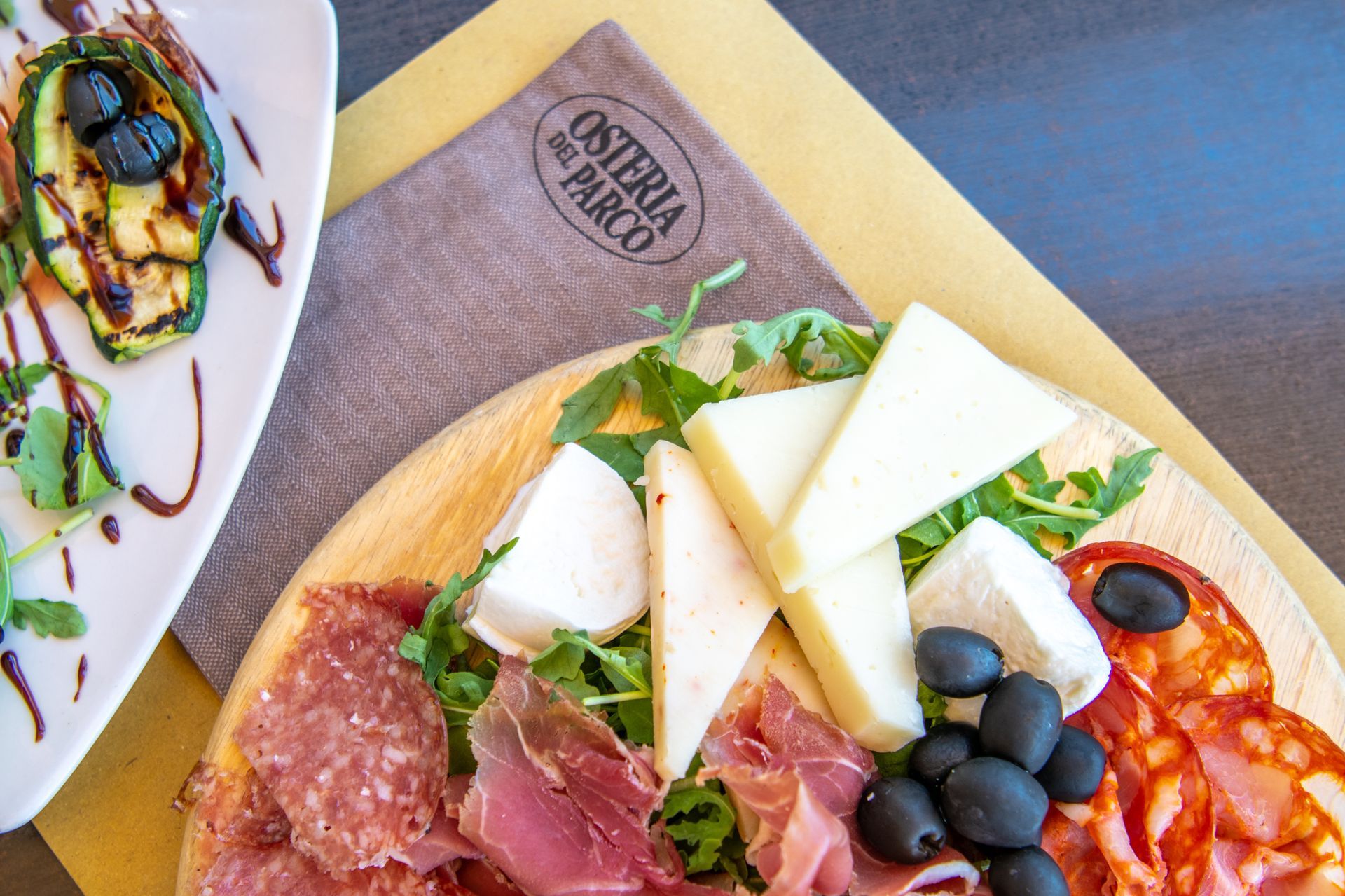 Tagliere di salumi con formaggi, olive, affettati e verdure su un vassoio di legno, accanto a un piattino da antipasto.
