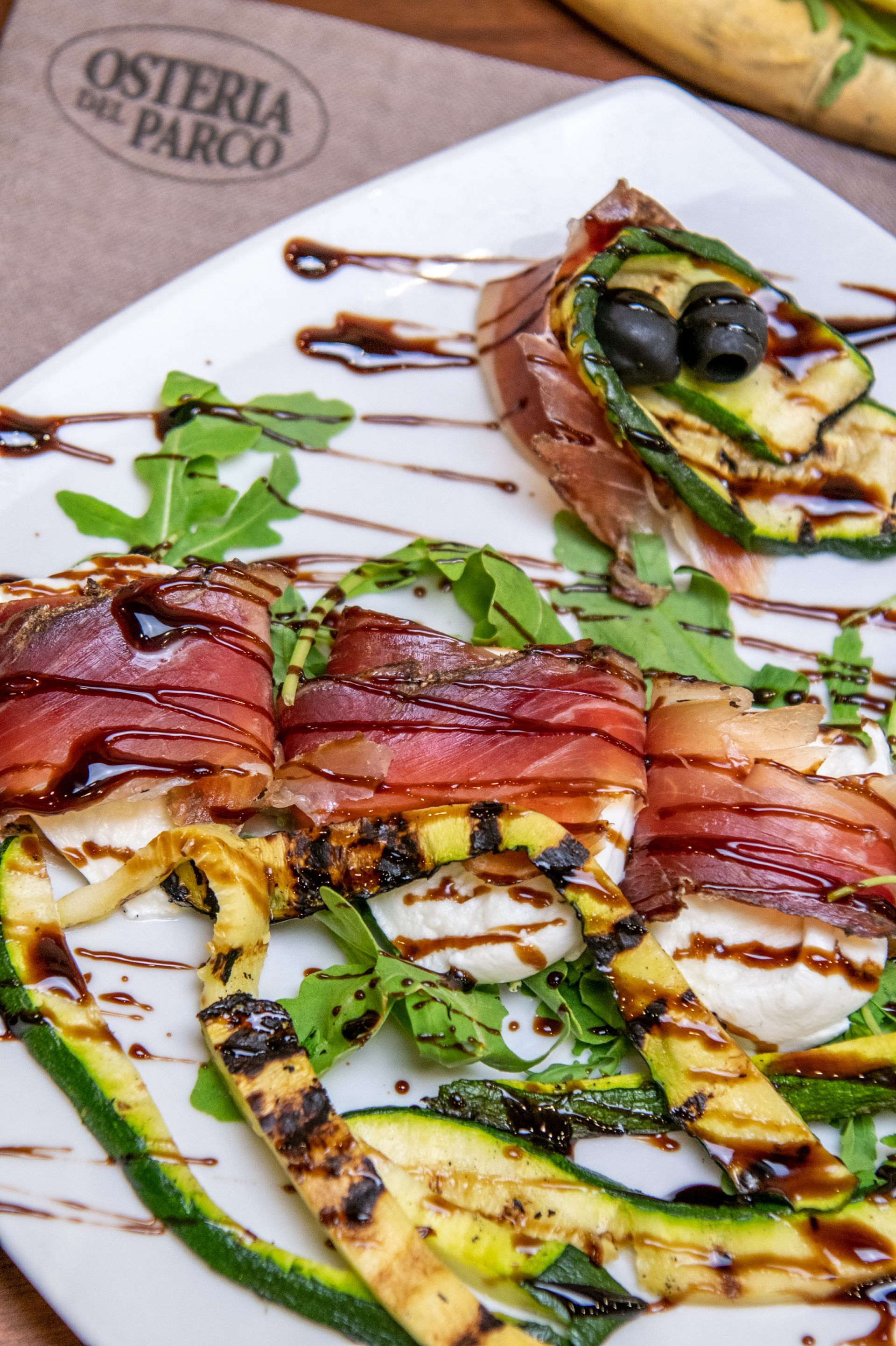 Piatto di verdure grigliate e prosciutto condito con salsa verde, servito su un piatto bianco.