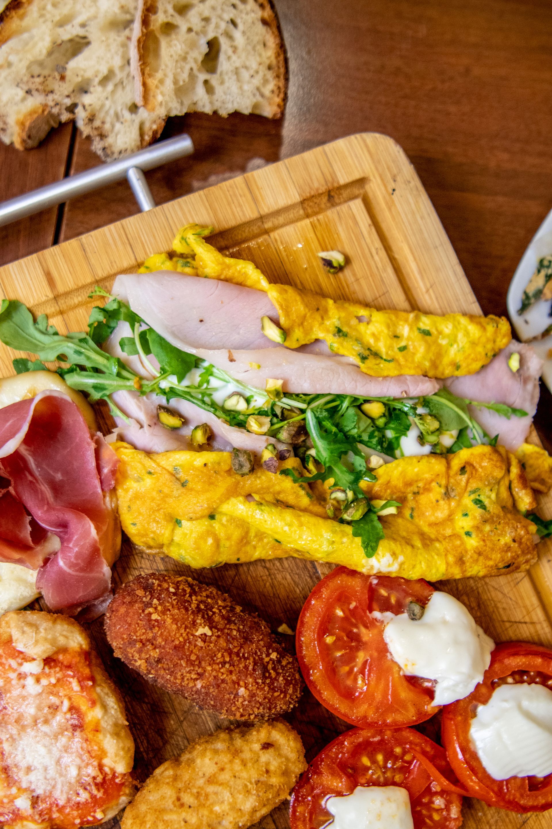Tagliere di salumi assortiti con pane, prosciutto crudo, fette di frittata, snack fritti e pomodori su un tagliere di legno.
