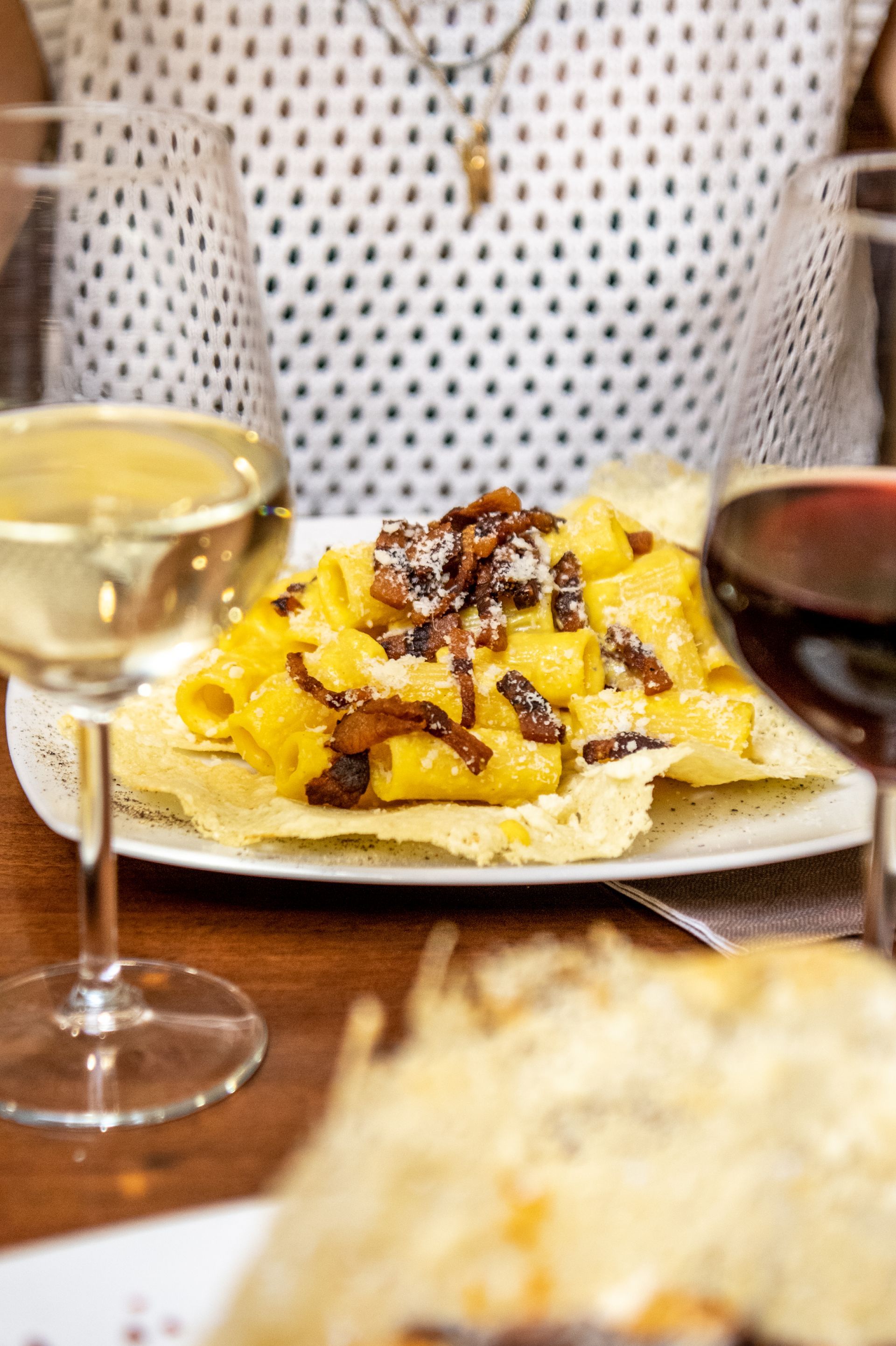 Una persona mangia una crêpe con frutta a un tavolo, con bicchieri di vino bianco e rosso nelle vicinanze.