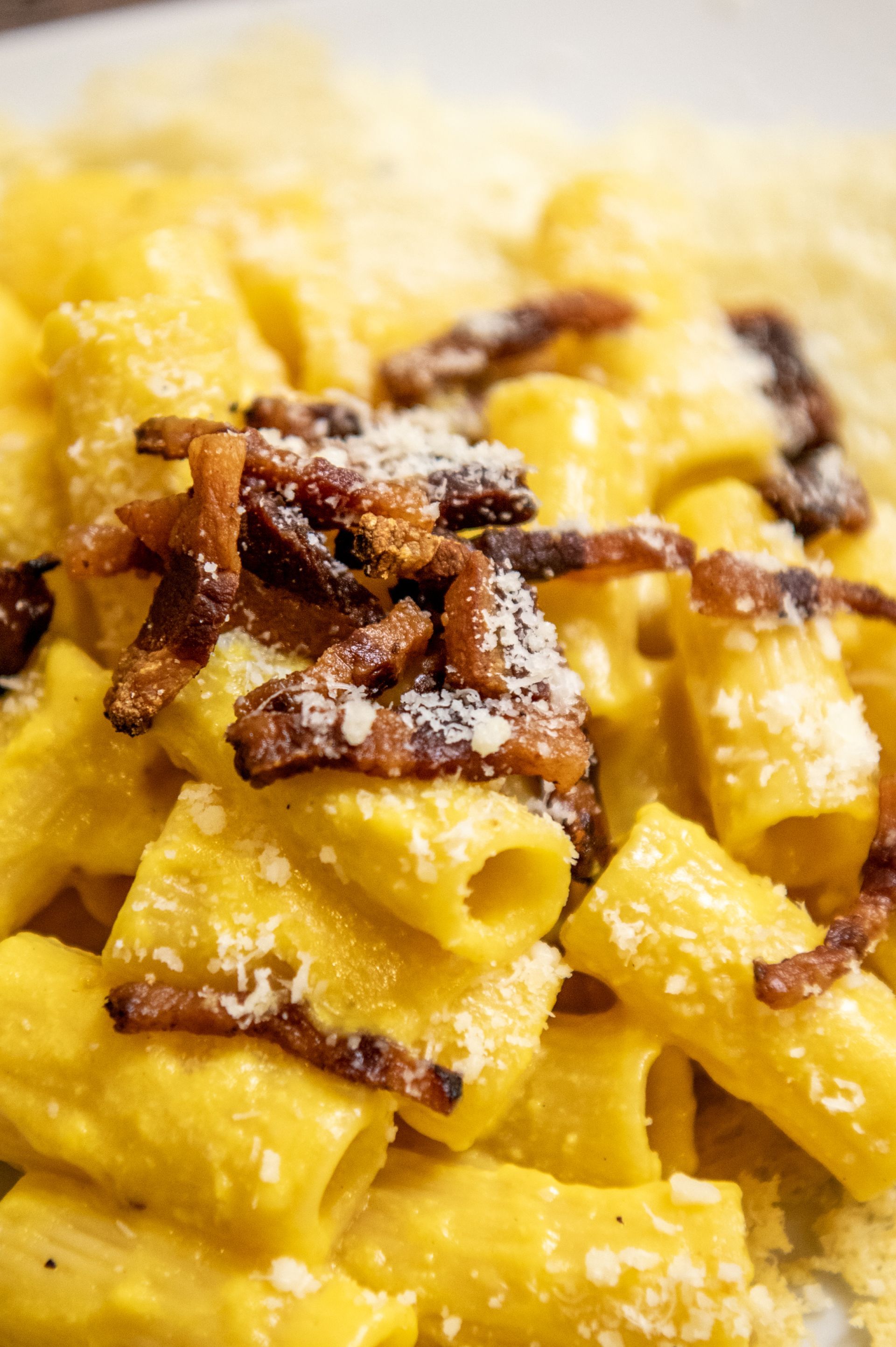 Primo piano di una cremosa pasta gialla con pezzetti di pancetta croccante e formaggio grattugiato.